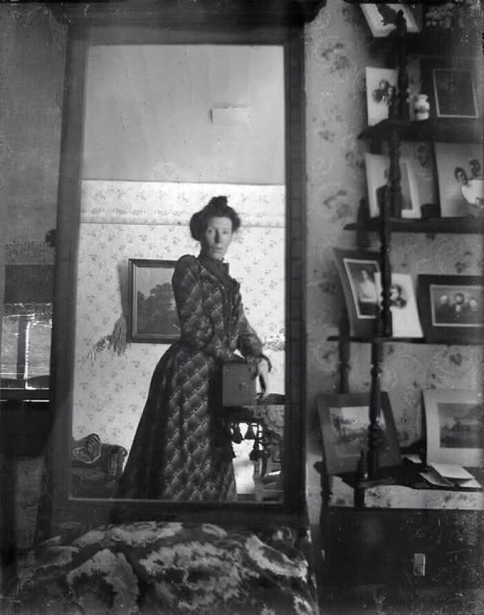 Victorian-Edwardian picture of a woman in period dress taking a self-portrait in a room filled with framed photos and wallpaper.