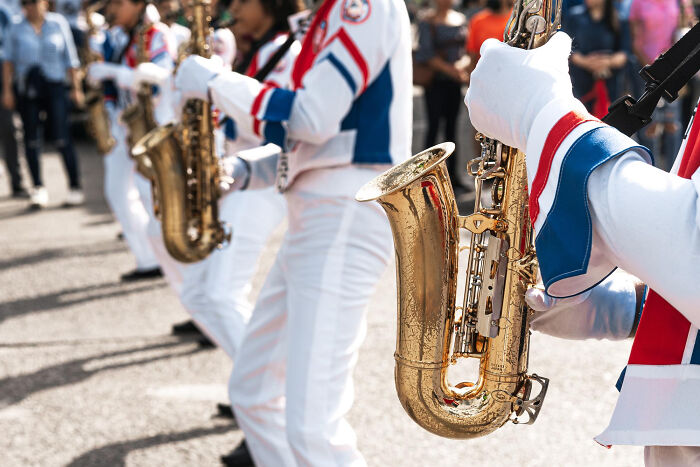 Marching band members playing saxophones in white uniforms during an outdoor event, illustrating stories of little lies backfiring.