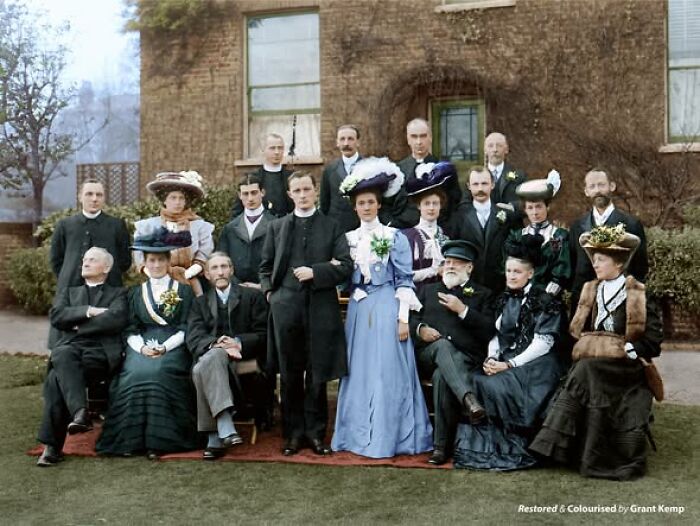 Group portrait of Victorian-Edwardian people dressed in period clothing posing outdoors in front of a brick building.