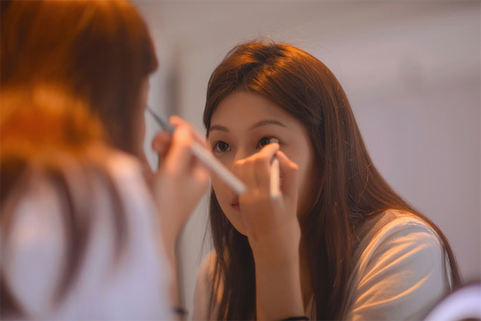 Woman applying makeup in front of mirror preparing for Friendsgiving amid dress-code betrayal controversy. Woman applying makeup in front of mirror preparing for Friendsgiving amid dress-code betrayal controversy.