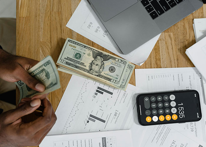 Hands counting cash on a desk with financial charts, calculator app, and a laptop showing terrifying things found online.
