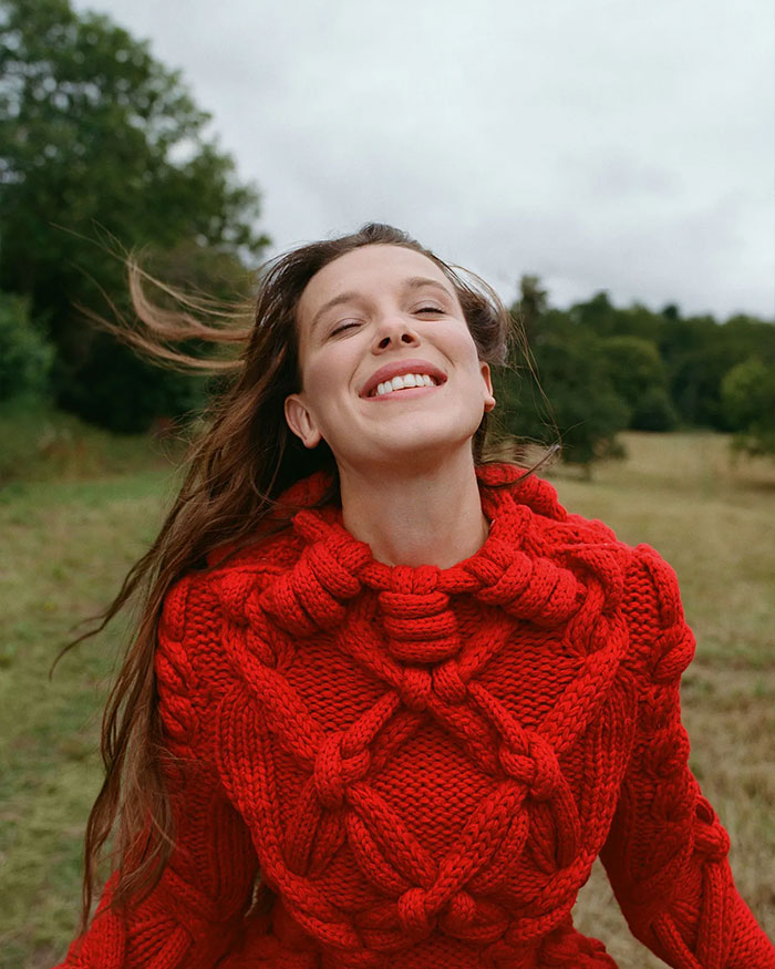 Young woman smiling outdoors wearing a chunky red knit sweater, symbolizing Millie Bobby Brown adopting a baby at 21. Young woman smiling outdoors wearing a chunky red knit sweater, symbolizing Millie Bobby Brown adopting a baby at 21.
