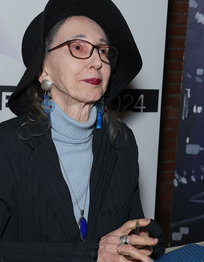 Joyce Carol Oates wearing a black hat and glasses, speaking at an event about Elon Musk controversy.
