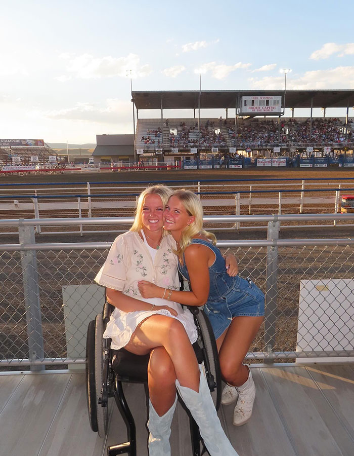 Girl who was paralyzed at 16 sitting in wheelchair with friend at outdoor event, smiling and enjoying the moment.