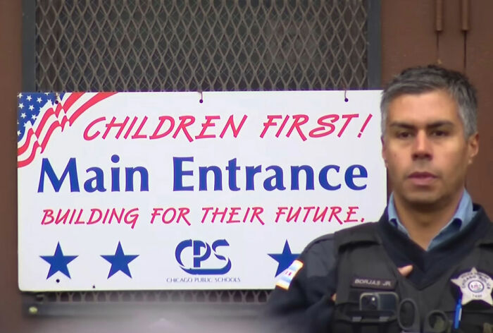 Police officer standing near Chicago Public Schools main entrance sign with children first message visible.