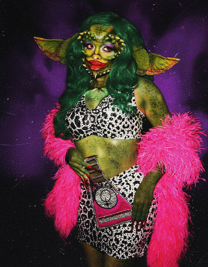 Celebrity wearing a detailed Halloween costume with green makeup, large ears, and a pink feathered jacket at a costume event.