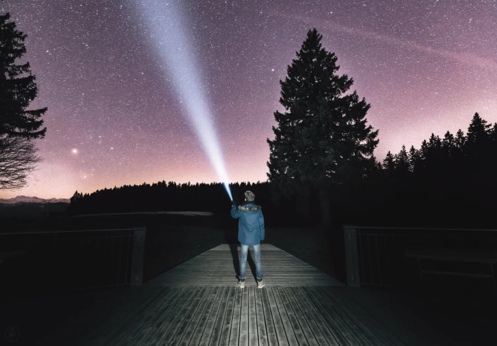 Person shining flashlight into starry night sky on wooden bridge, evoking best scary games to play with friends IRL.