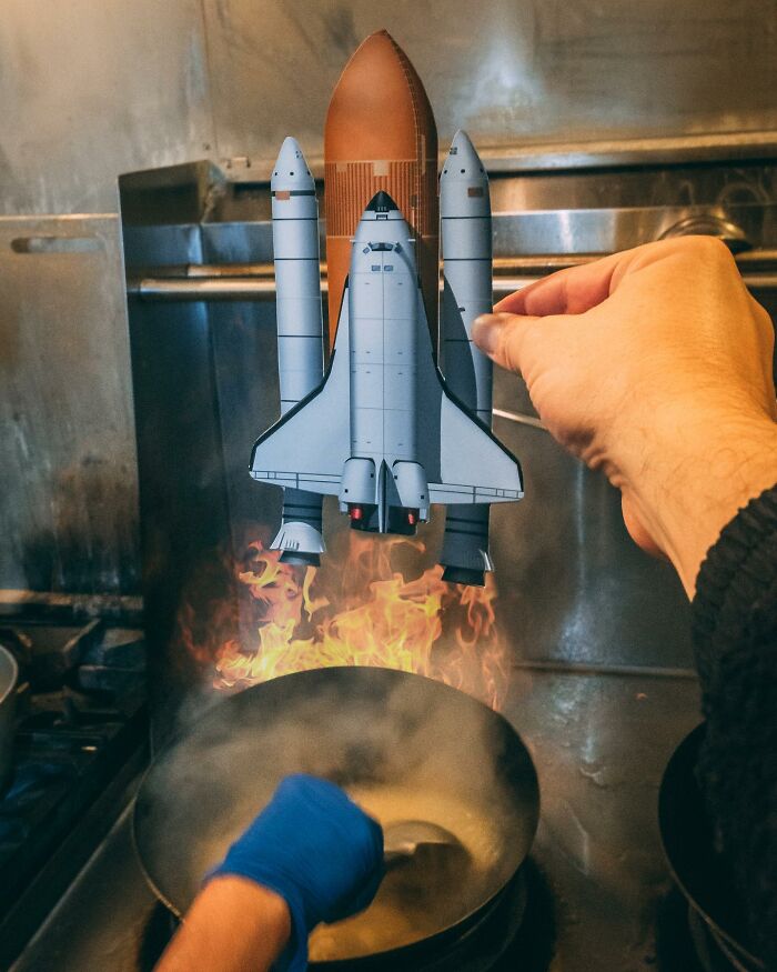 Hand holding a paper cutout of a space shuttle with flames, interacting with real life kitchen fire and cooking.