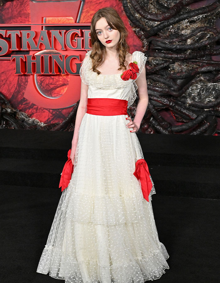 Young woman in white dress with red accents poses at Stranger Things 5 red carpet premiere, showcasing strange fashion choices.