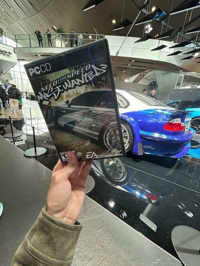 Hand holding a Need for Speed Most Wanted game case with a blue sports car displayed in the background in a showroom.
