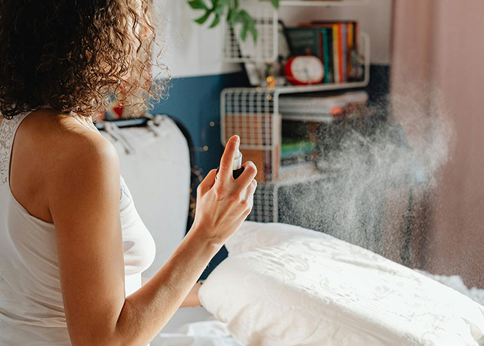 Woman spraying cleaning solution on a pillow in a bedroom applying genius cleaning hacks inspired by laziness.