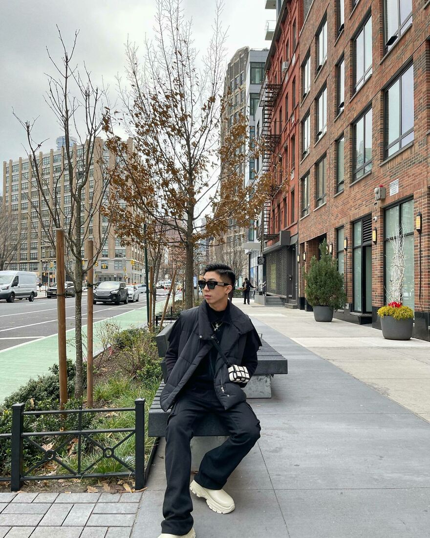 Young Asian man sitting on a bench in urban street setting, dressed in black, challenging Asian stereotypes in modern world.
