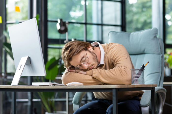 Young man asleep at desk in modern office, symbolizing realization of compulsory things in life being optional.