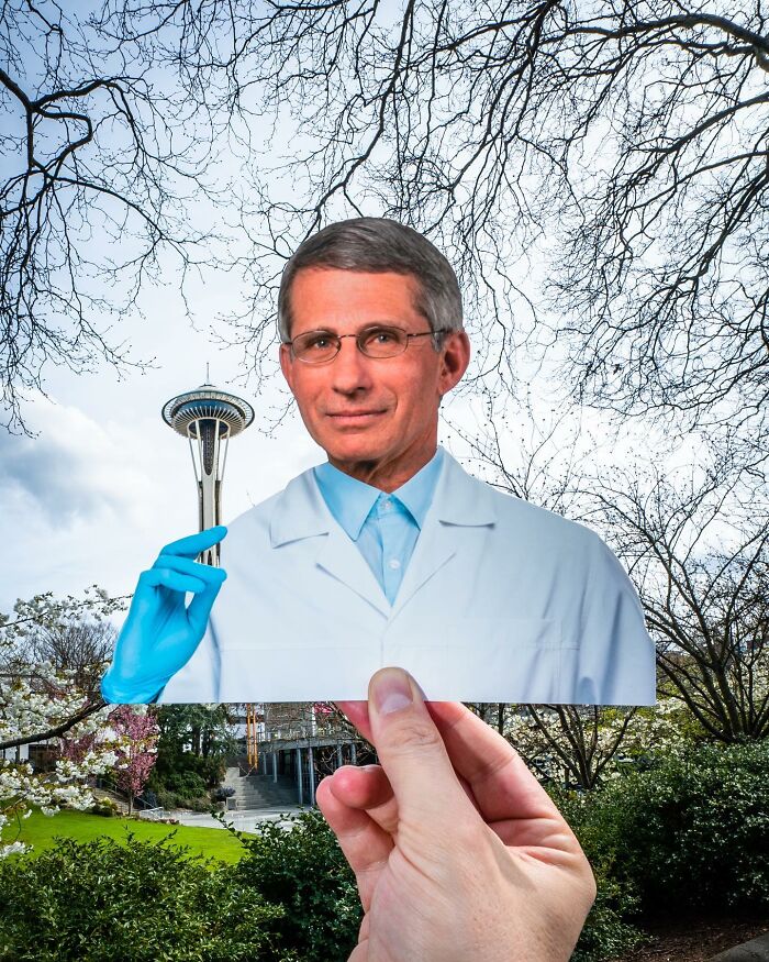 Hand holding a paper cutout of a man with a blue glove interacting with the Seattle Space Needle outdoors.