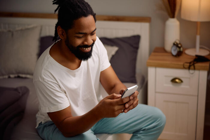 Man smiling discreetly at phone while sitting on a bed, hinting at subtle signs of cheating behavior.