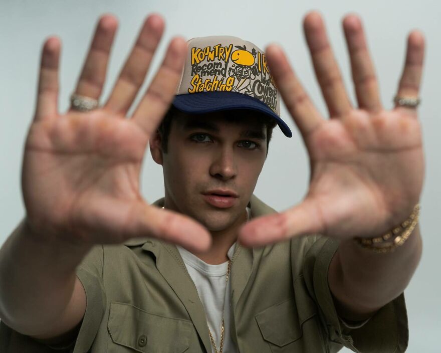 Austin Mahone wearing a graphic cap and casual outfit, posing with hands extended, related to celebrities on fan pages.