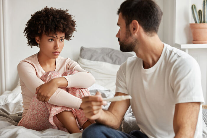 Couple sitting on bed as clueless dad holds pregnancy test while wife looks concerned in a home setting.