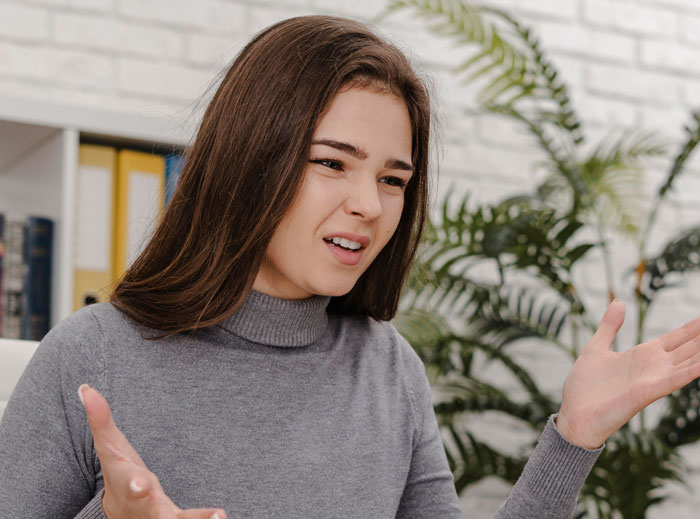 Young woman in gray sweater expressing frustration indoors, illustrating bride wanting to upgrade honeymoon at bridesmaid&rsquo;s expense concept.