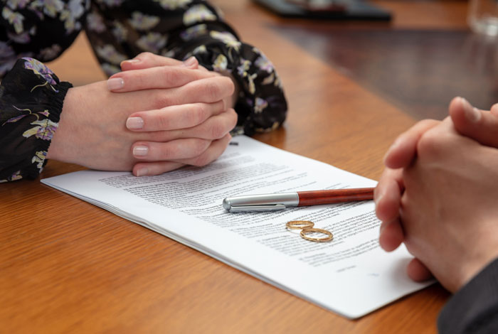 Two people with folded hands sitting at a table with a document, pen, and wedding rings symbolizing family drama.