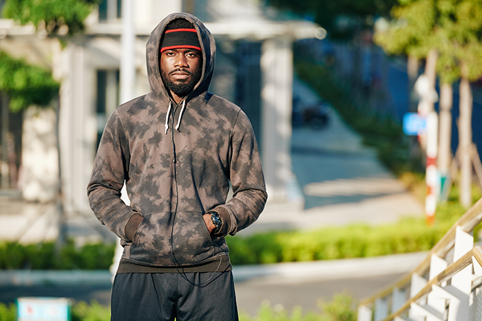 Young man in a hoodie standing outdoors with a serious expression, representing tension in a petty neighbor conflict scenario.