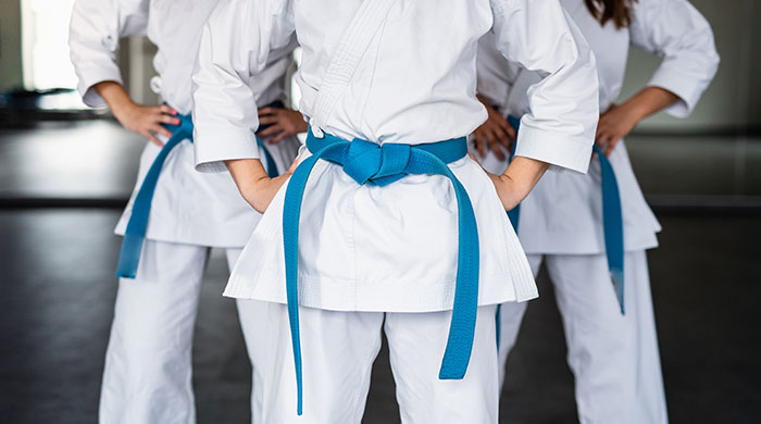 Three teens wearing white martial arts uniforms with blue belts, standing with hands on hips in a dojo.