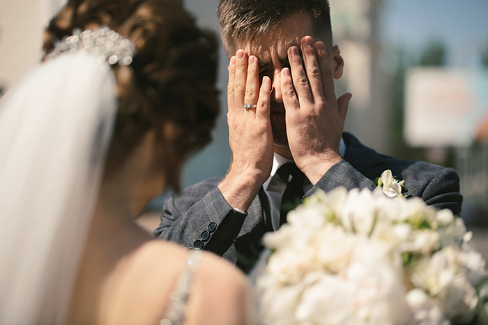 Groom covering face in distress during wedding ceremony as bride stands nearby holding bouquet at outdoor altar.