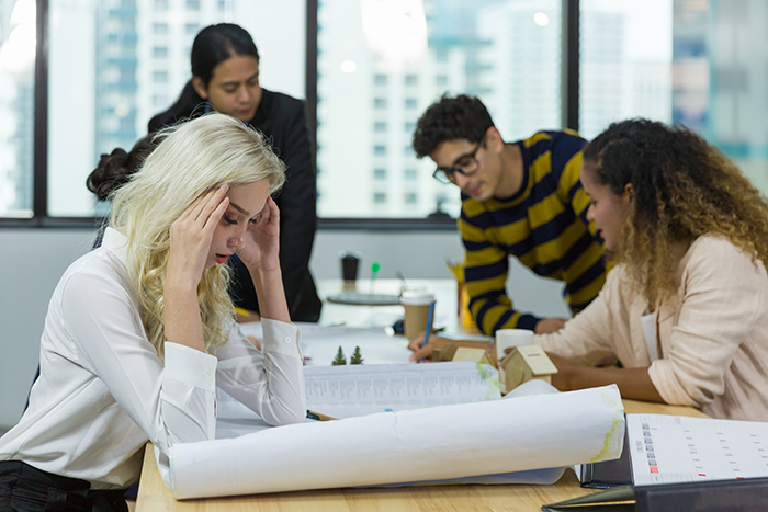 Stressed employee overwhelmed by multitasking the roles of four people while colleagues discuss work in a busy office.