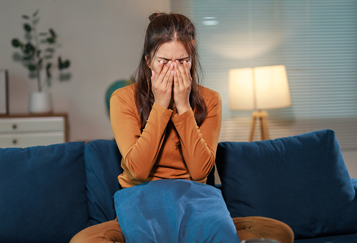 Woman in a mustard sweater sitting on a blue couch, covering her face, reflecting confusion about wedding and marriage.