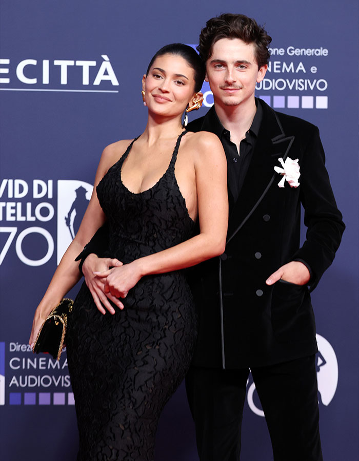 Timothée Chalamet in a black suit with a white flower and a woman in a black dress at a red carpet event. Timothée Chalamet in a black suit with a white flower and a woman in a black dress at a red carpet event.