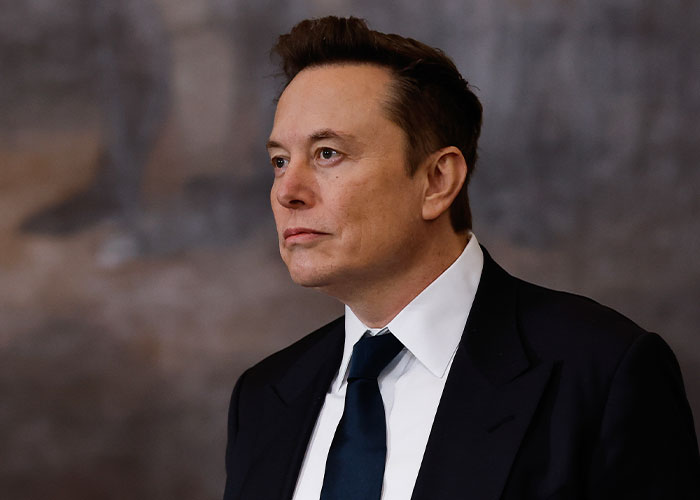 Elon Musk in a dark suit and tie, looking serious during a formal event with a blurred background.