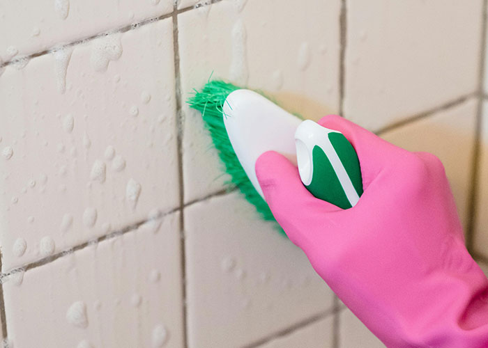 Hand in pink glove using green brush to scrub tiled wall with soap, illustrating genius cleaning hacks inspired by laziness.