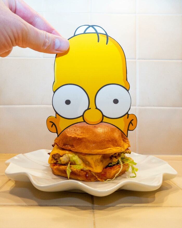 Hand holding a brilliant paper cutout of a cartoon face perfectly interacting with a cheeseburger on a plate.