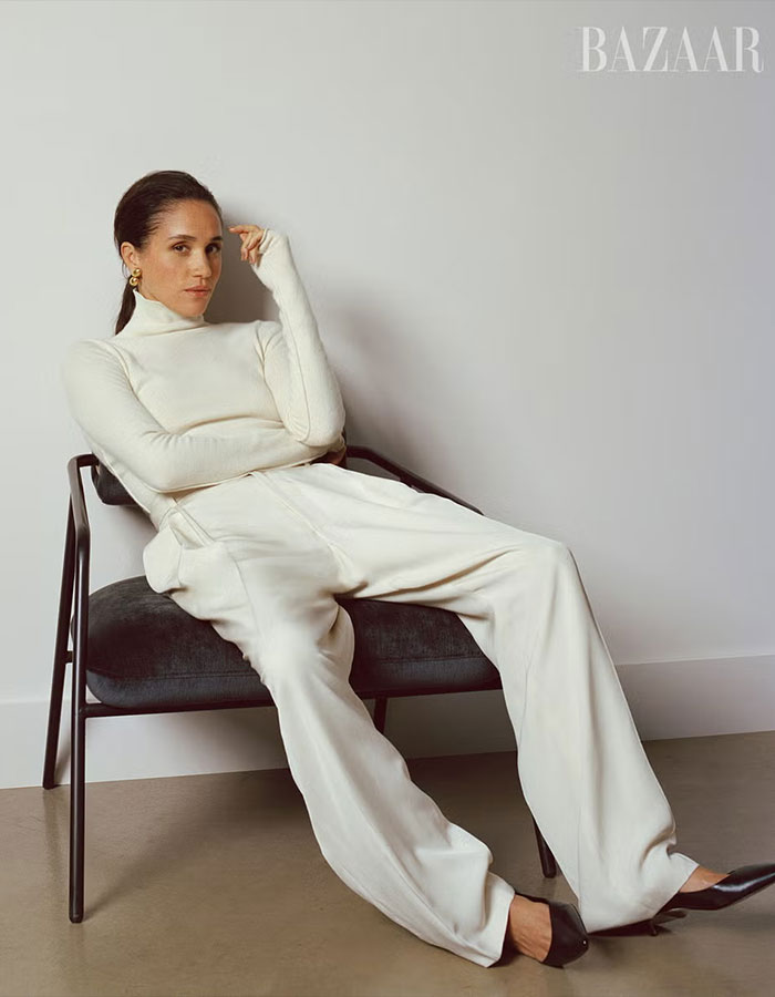 Woman in white turtleneck and trousers seated on chair, showcasing Meghan Markle Harper&rsquo;s Bazaar moment style and elegance.