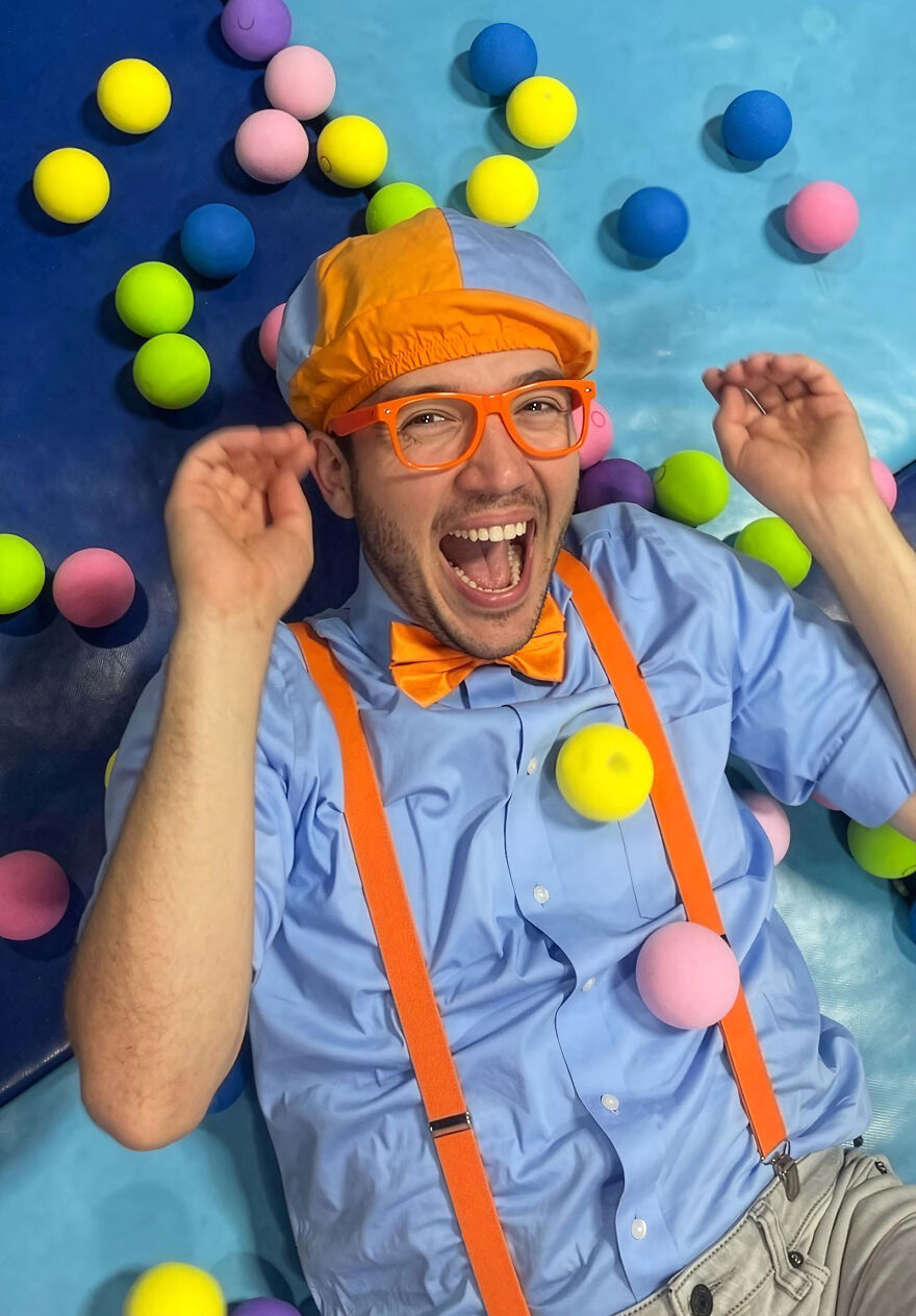 A Blippi wearing orange suspenders and bow tie, colorful hat and glasses, smiling among yellow, pink, blue, and green balls related to Blippis.