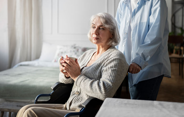 Elderly woman in a wheelchair holding a cup, appearing thoughtful, with a caregiver standing behind her at home.