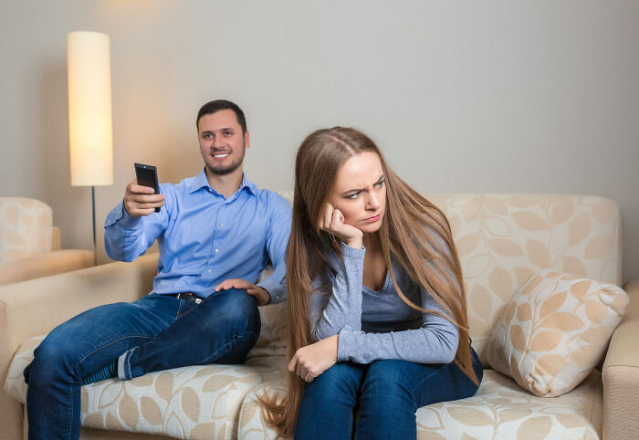 A couple sitting on a couch, the man smiling and holding a remote while the woman looks upset, depicting personal lessons.