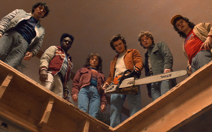 Stranger Things cast members looking downwards with one holding a chainsaw in a suspenseful scene.
