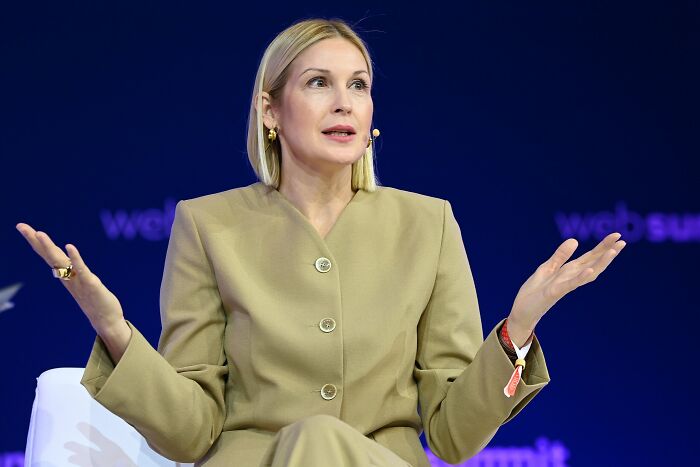 Blonde woman in beige suit speaking on stage, exemplifying style icons whose looks changed fashion forever.