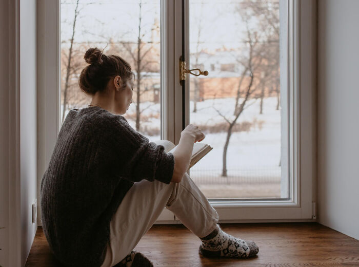 Young woman sitting on the floor by the window reading a book, illustrating signs child never loved properly concept.