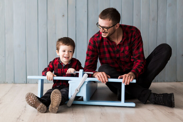 Father and son wearing matching red plaid shirts playing together with a blue wooden chair inside a room.