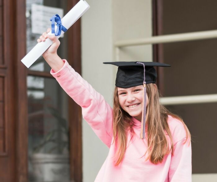 Smiling young graduate in cap and pink sweatshirt holding diploma celebrating achievements outdoors.