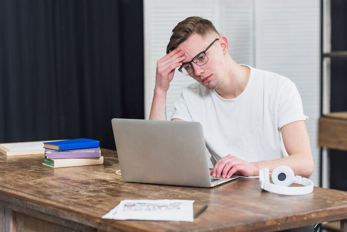 Young man with glasses looking stressed while working on a laptop, reflecting on a little lie that backfired hard