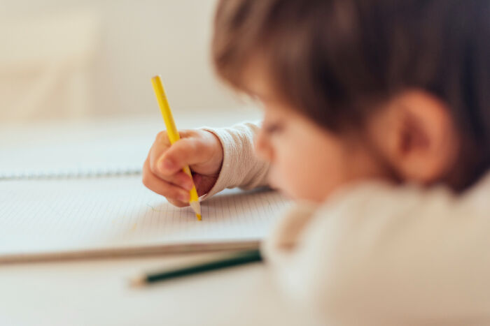 Child writing with a yellow pencil in a notebook, showing kids doing something weird clearly passed down from parents.