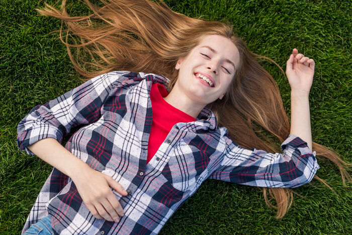 Young woman lying on green grass, smiling with eyes closed, capturing a bizarre moment that feels strangely sane.