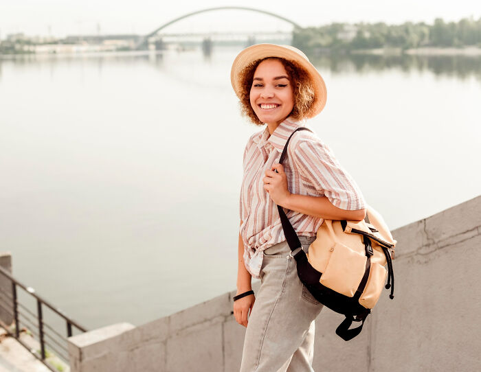 Young woman smiling by the water, wearing a hat and backpack, symbolizing people who disappeared to start new lives.