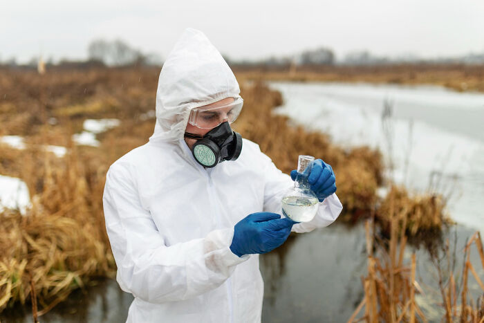 Scientist in protective gear testing water quality in an outdoor wetland, illustrating environmental research challenges.
