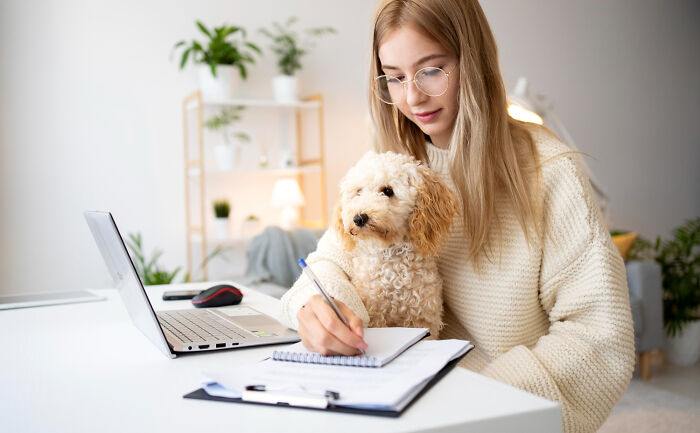A dog mom working from home with her curly dog sitting on her lap while she writes notes at a desk.