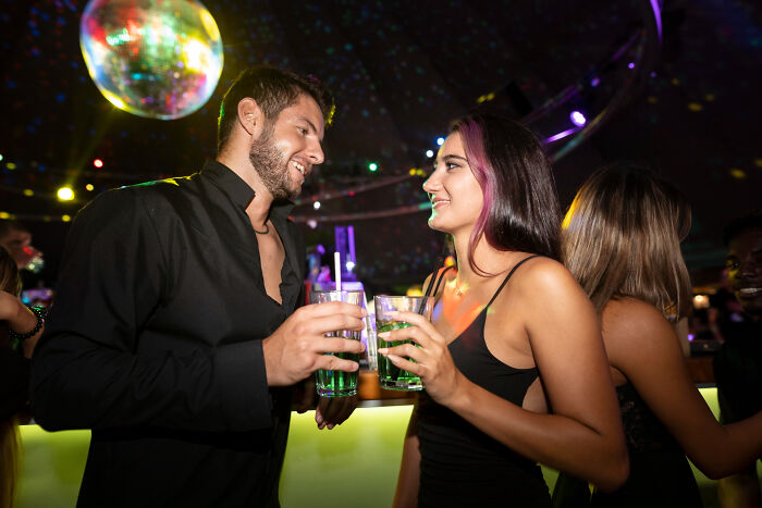 Two people in a nightclub holding drinks and smiling, illustrating social situations where little lies backfired.
