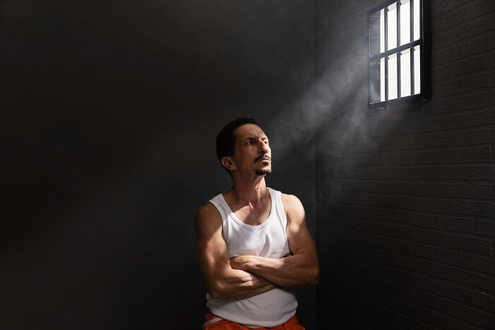 Man in prison uniform looking thoughtfully toward barred window with light shining, illustrating comically absurd facts concept.