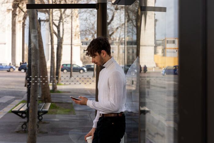 Young man standing at a bus stop looking at his phone, illustrating people open up about little lies that backfired hard.